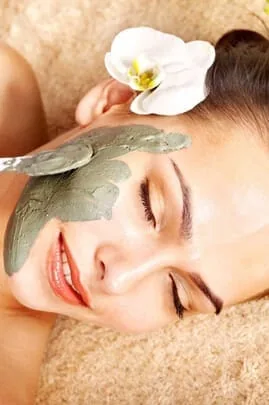 Woman relaxing with a green herbal clay face mask and a white orchid in her hair during a natural facial