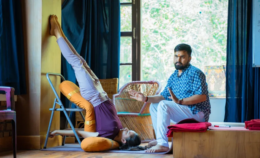 Teacher guiding a student in a chair-supported shoulderstand during yoga teacher training