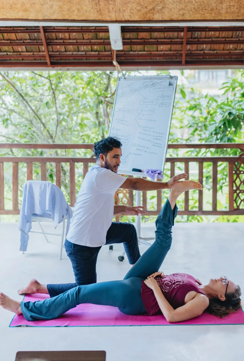 A teacher assisting a student in a supine leg stretch in the shala