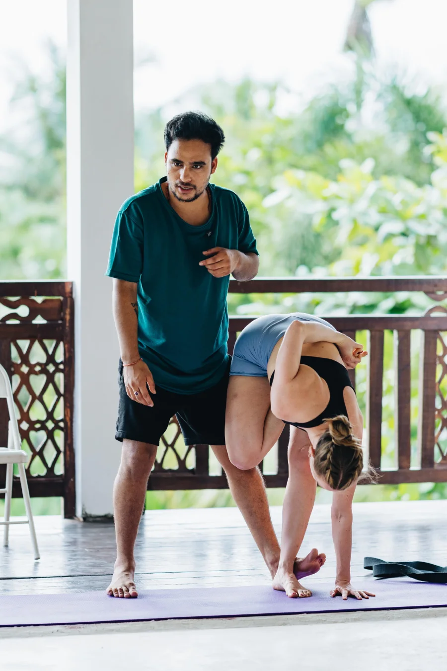 A teacher guiding a student in a standing forward bend with a bind