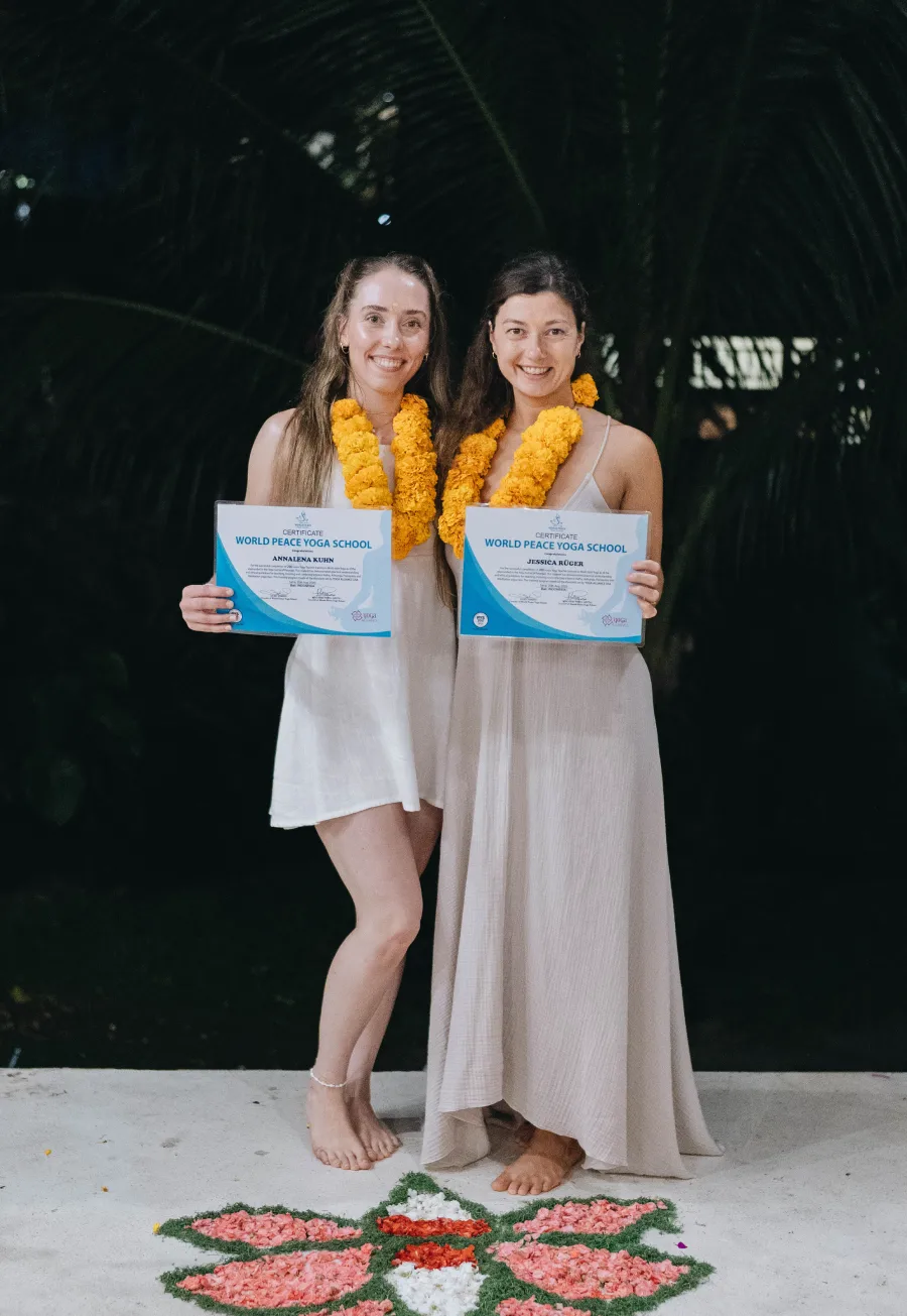 Two graduates in marigold garlands holding their World Peace Yoga School certificates