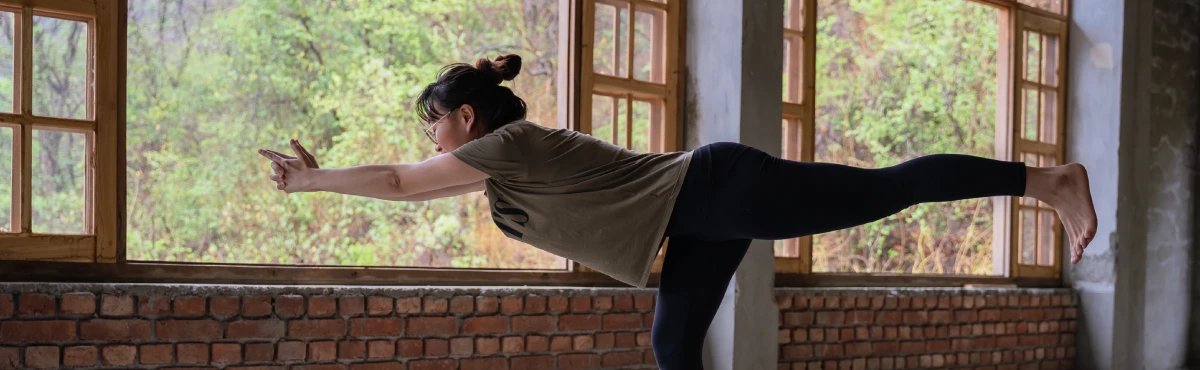 A student balancing in Warrior III by the yoga hall windows