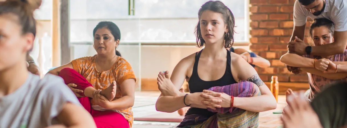 Students in a seated hip-opening pose while a teacher gives an adjustment