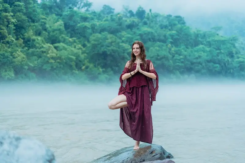 A student standing in tree pose on a rock beside the misty Ganga