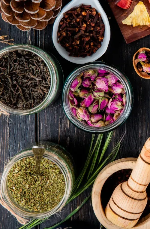 Jars of dried herbs, rose buds and spices beside a wooden pestle