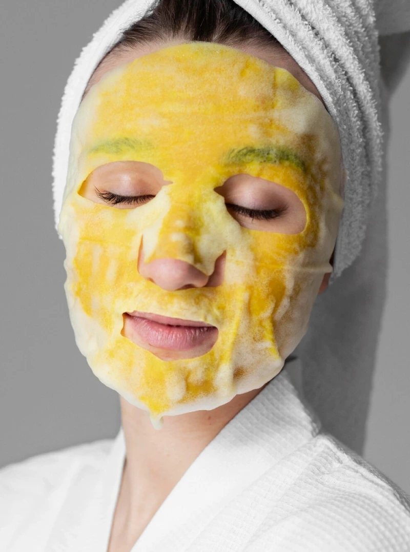 A relaxed woman in a towel wrap with a turmeric sheet mask on her face