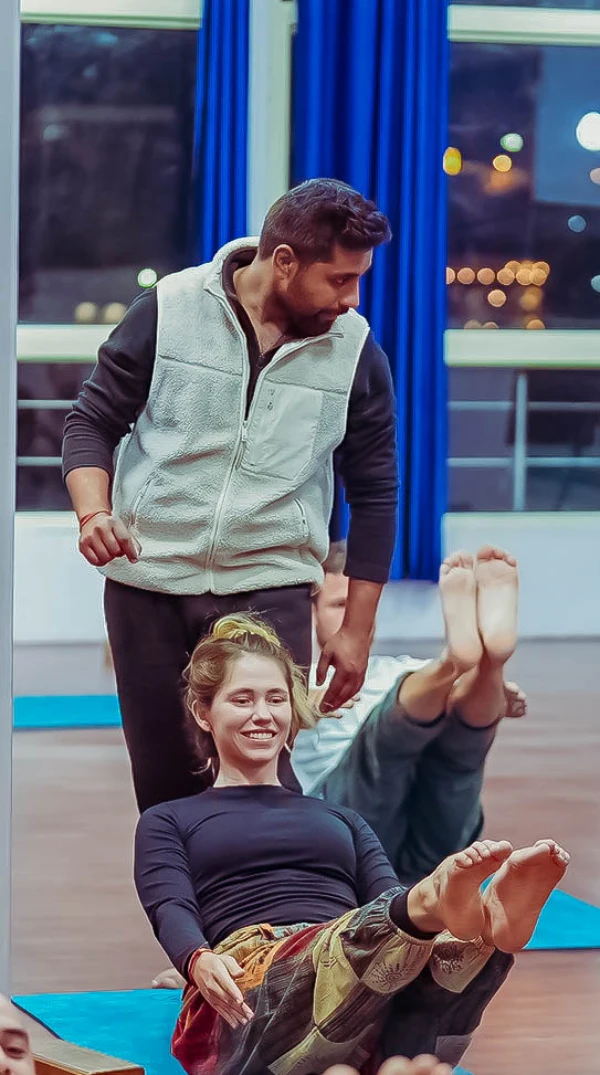 A teacher guiding a smiling student through a boat pose in an evening class