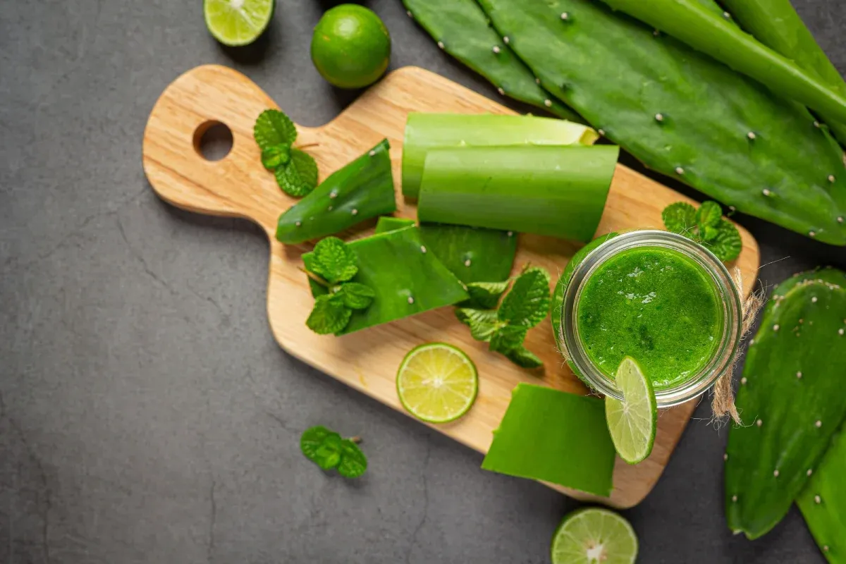 Fresh aloe vera, cactus leaves, limes, and a green juice on a wooden board