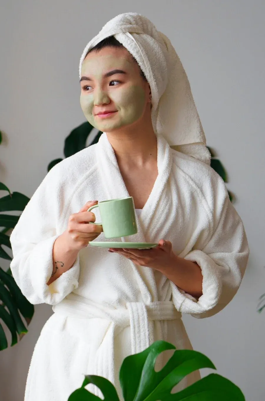 A woman in a bathrobe and green face mask holding a cup of tea