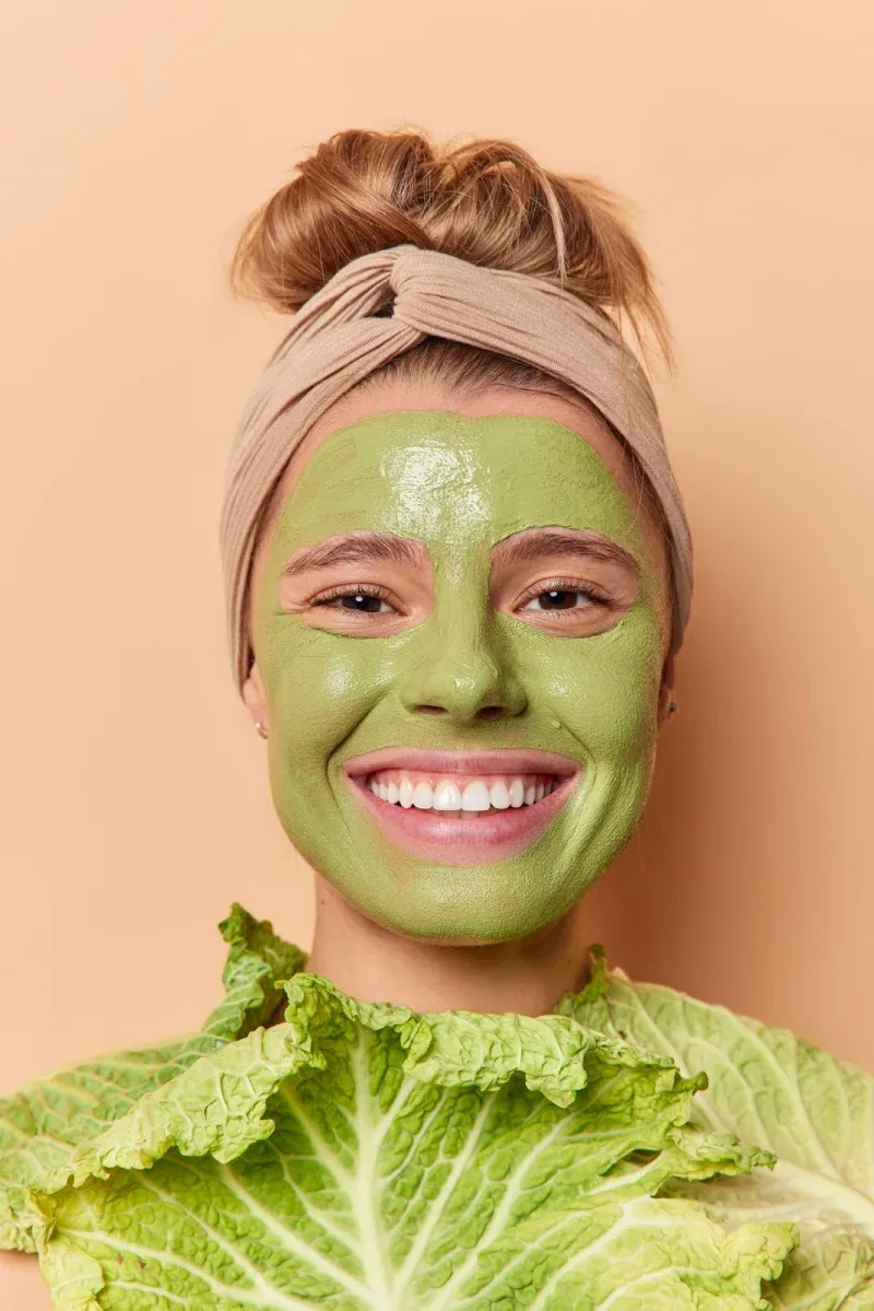 A laughing woman in a green face mask behind fresh cabbage leaves