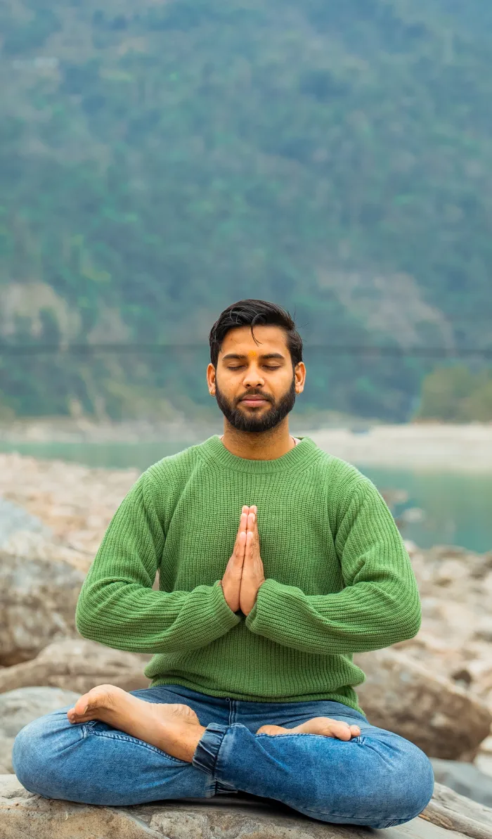 A man in a green sweater meditating with palms joined on a riverside rock