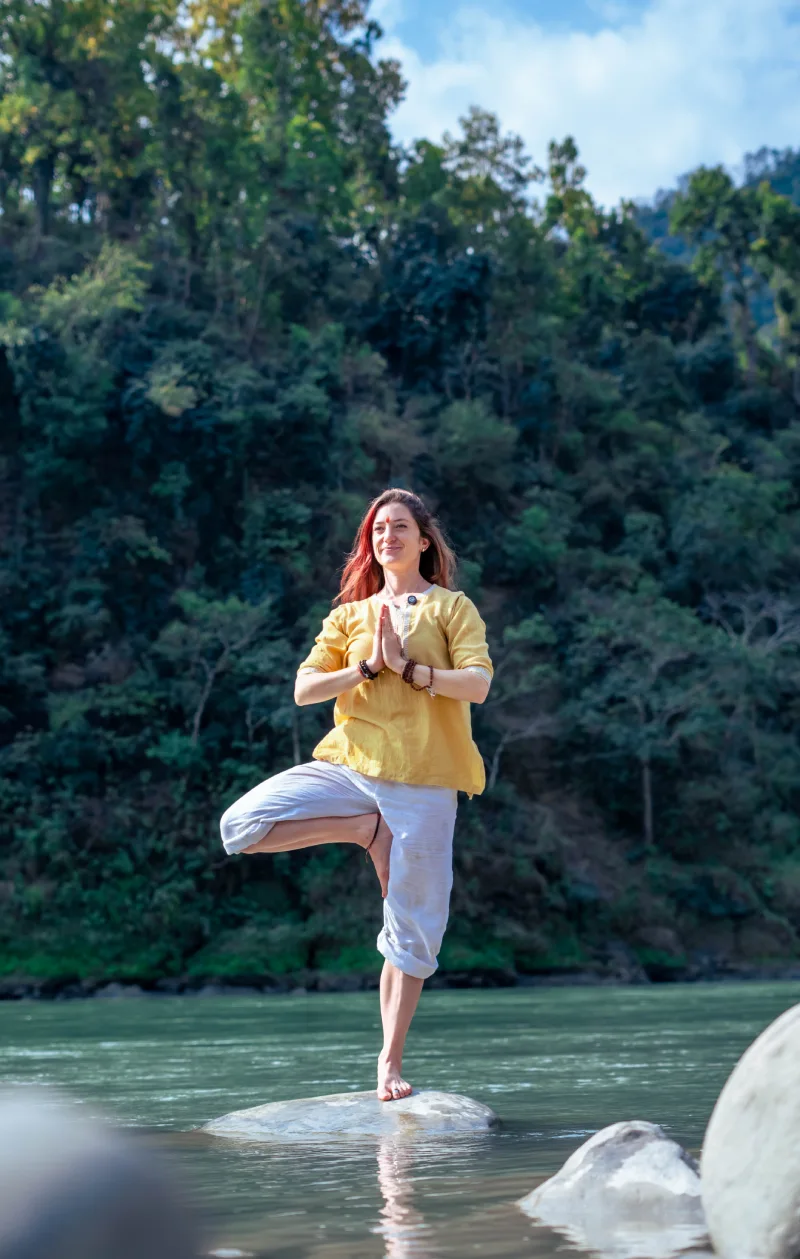 A smiling woman in tree pose on a rock in the Ganges