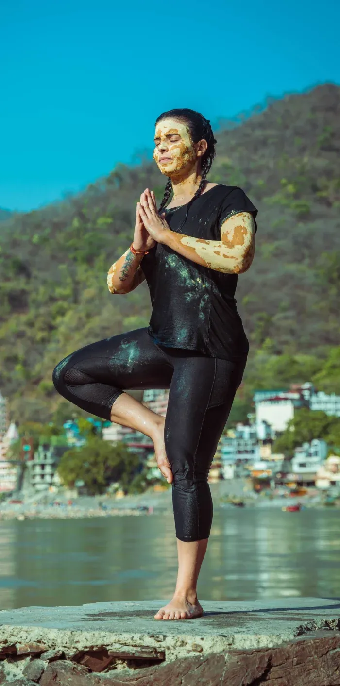 A woman in healing mud balancing in tree pose on a riverside rock