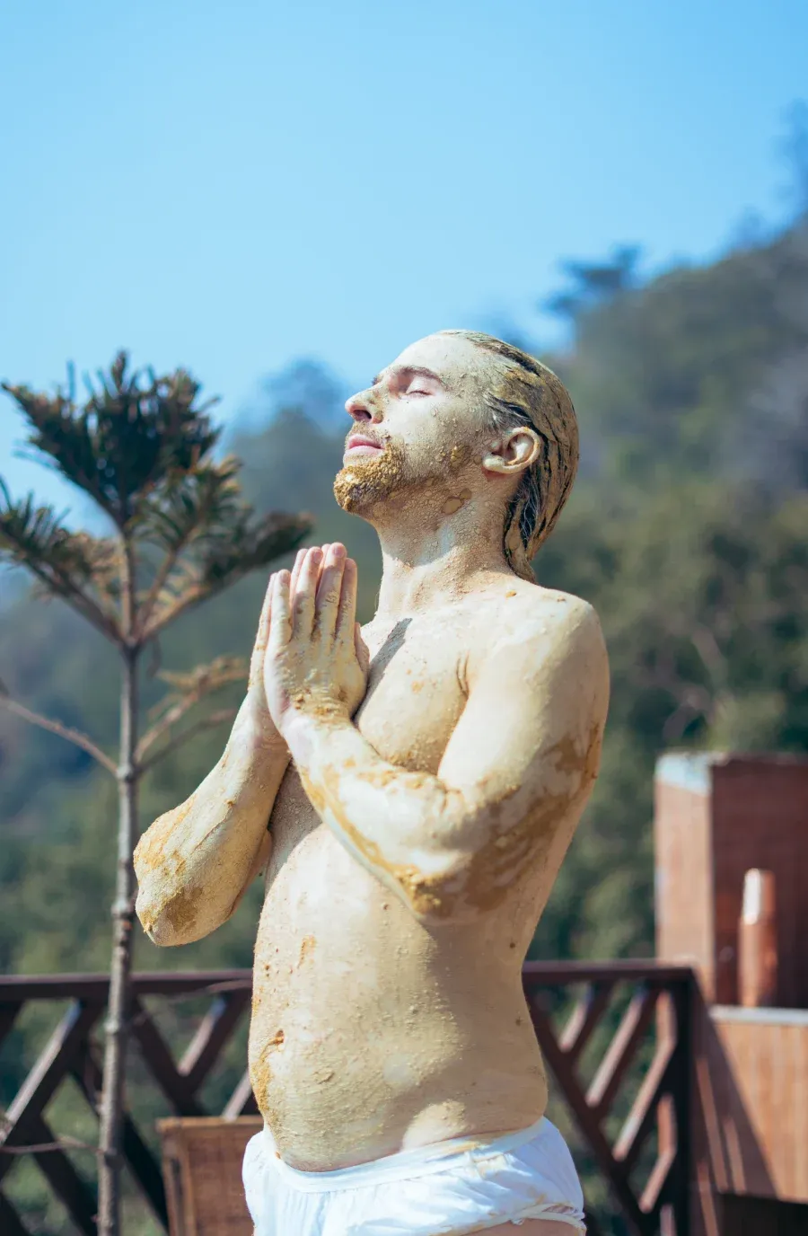 A man covered in clay standing with folded hands in the sun