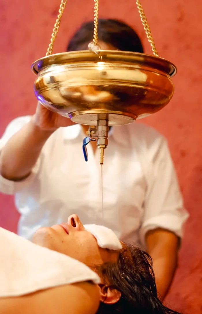 Warm oil flowing from a brass vessel onto a woman’s forehead during Shirodhara