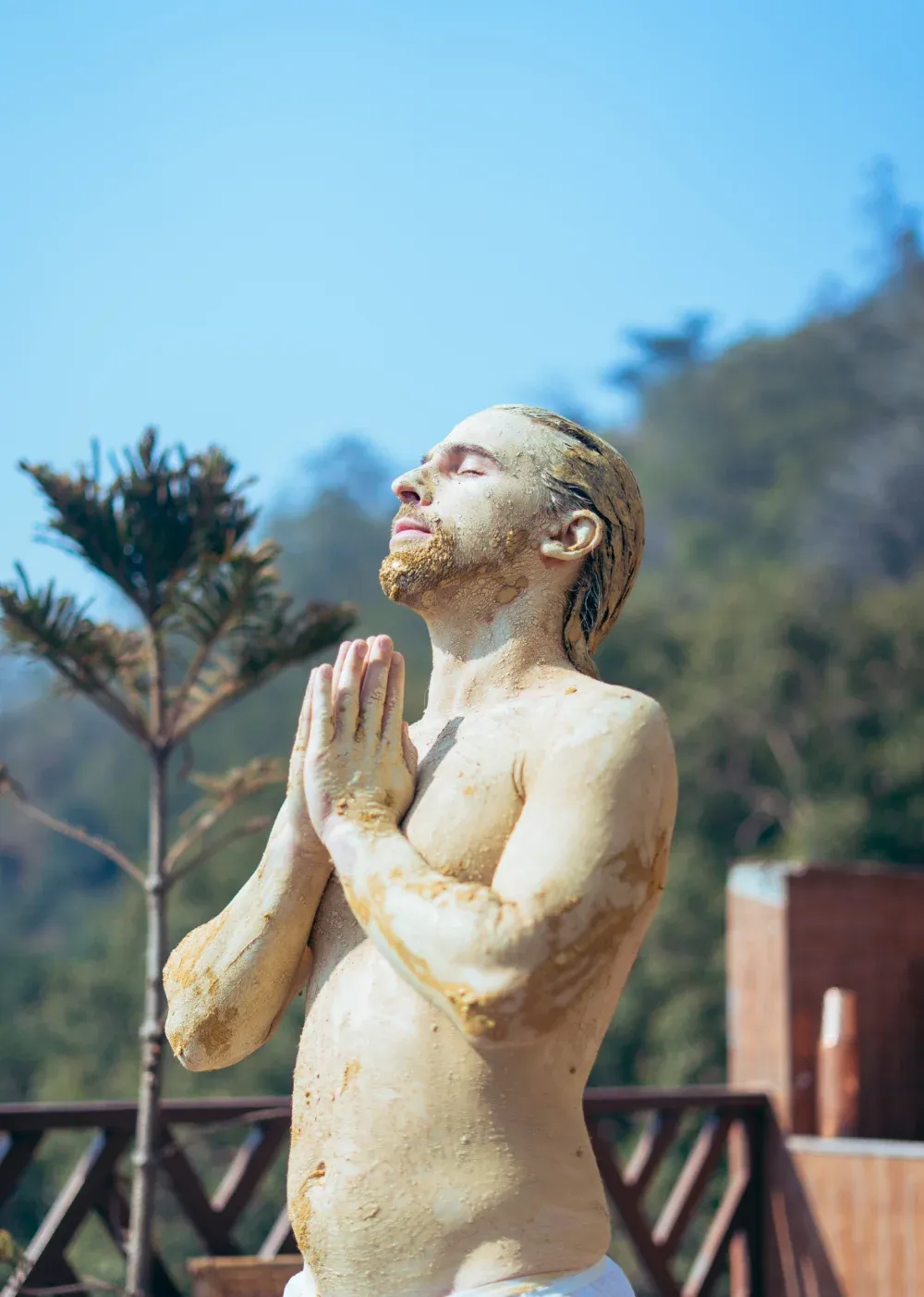 A man covered in healing clay standing with folded hands in the sun