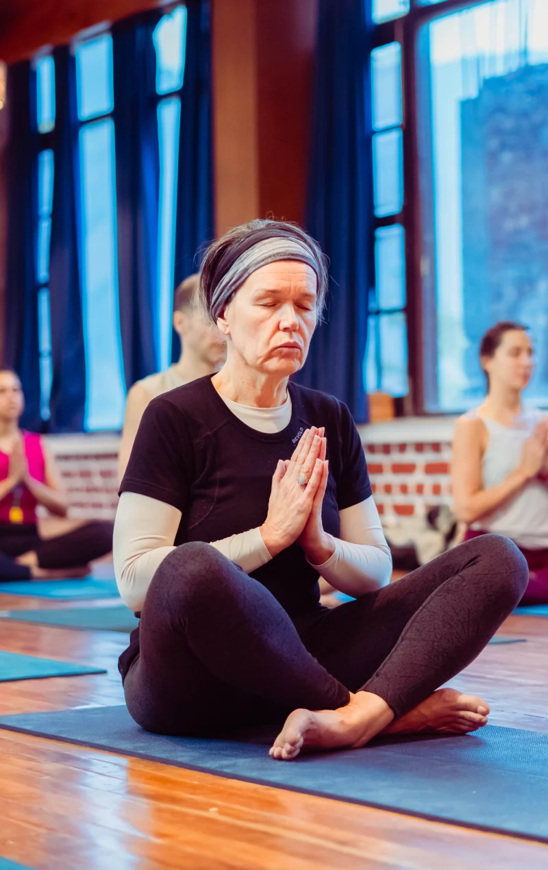 An older woman meditating cross-legged with palms joined in a yoga hall