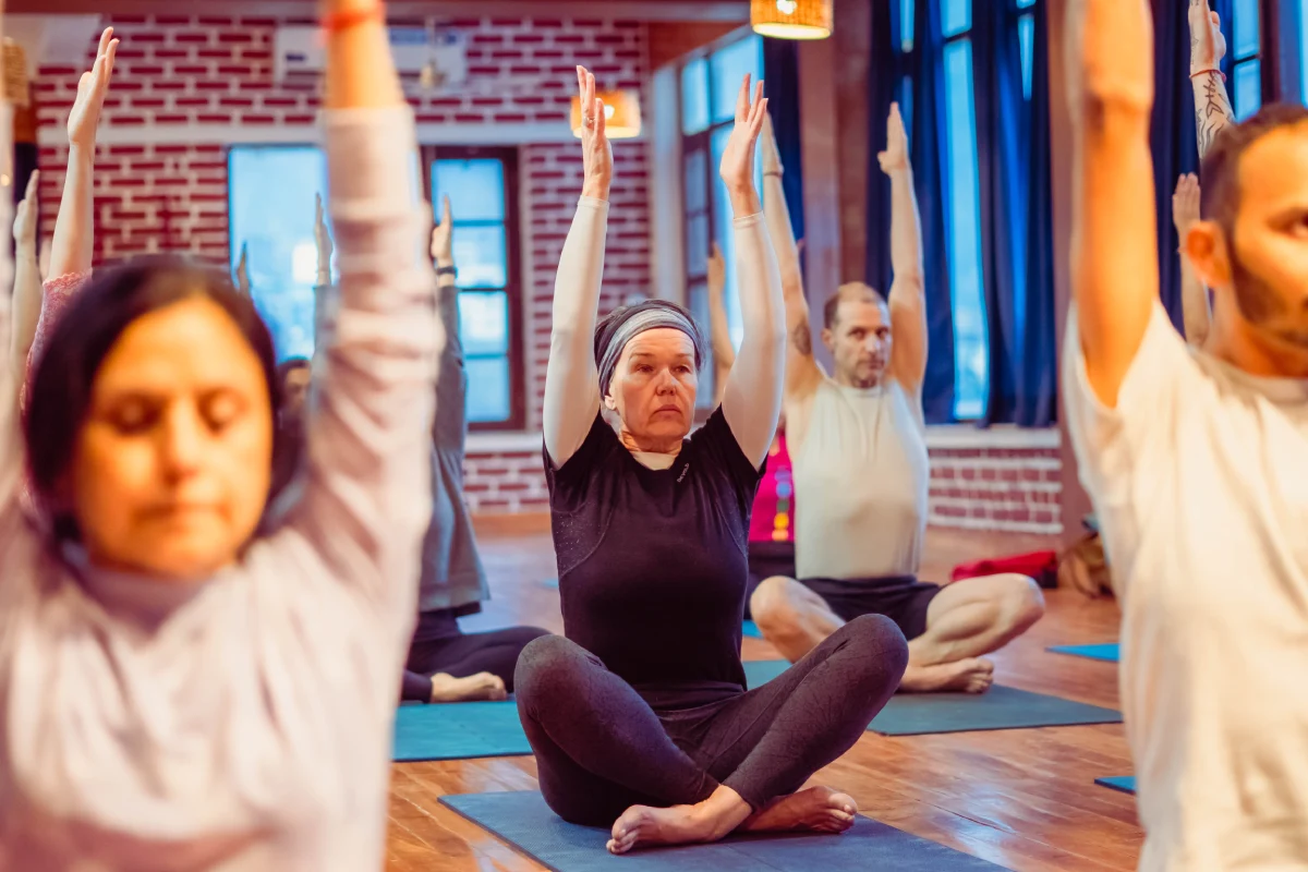 An older woman seated with arms raised among a group in a yoga hall