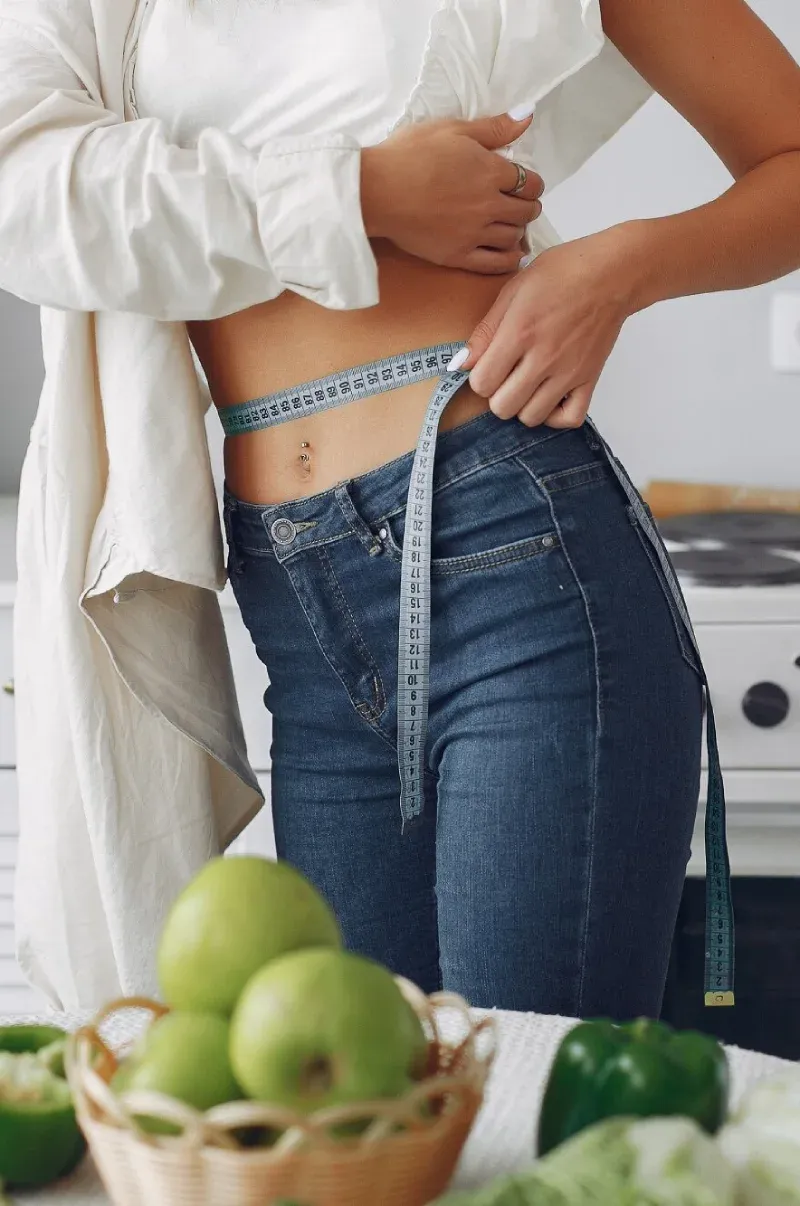 A woman measuring her waist beside a basket of green apples