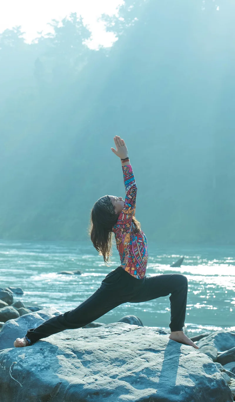 A woman in a crescent lunge on a rock beside the Ganges in morning light