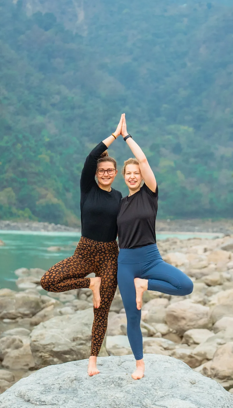 Two smiling women in tree pose with palms joined overhead by the river