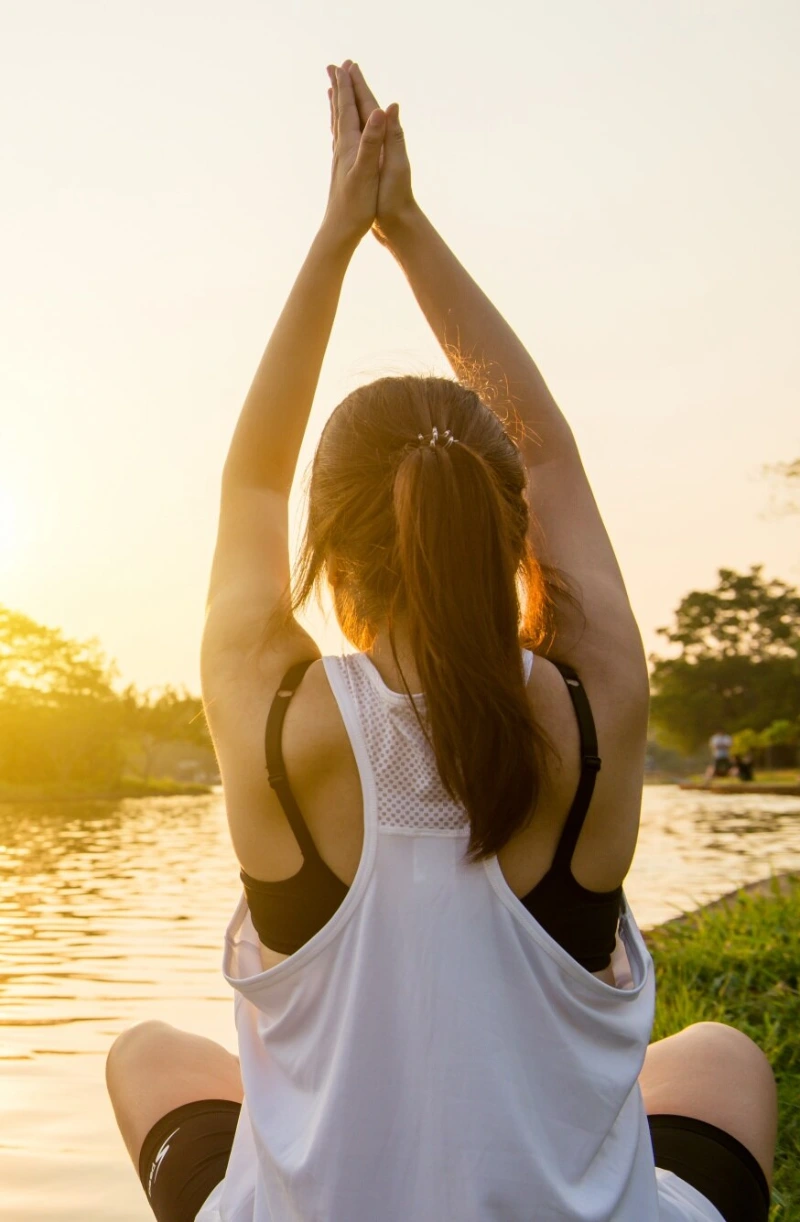 A woman seated by a lake at sunset with palms joined overhead
