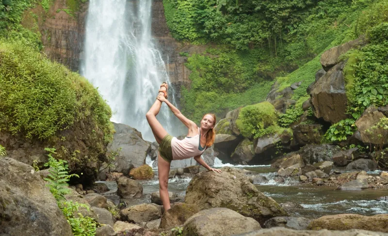 A student in a standing split on the rocks in front of a jungle waterfall
