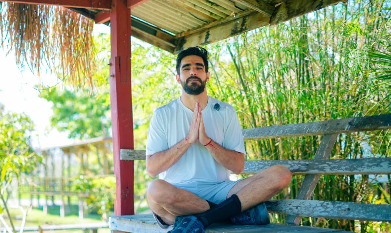 A student sitting cross-legged with hands in prayer under a thatched shelter beside bamboo